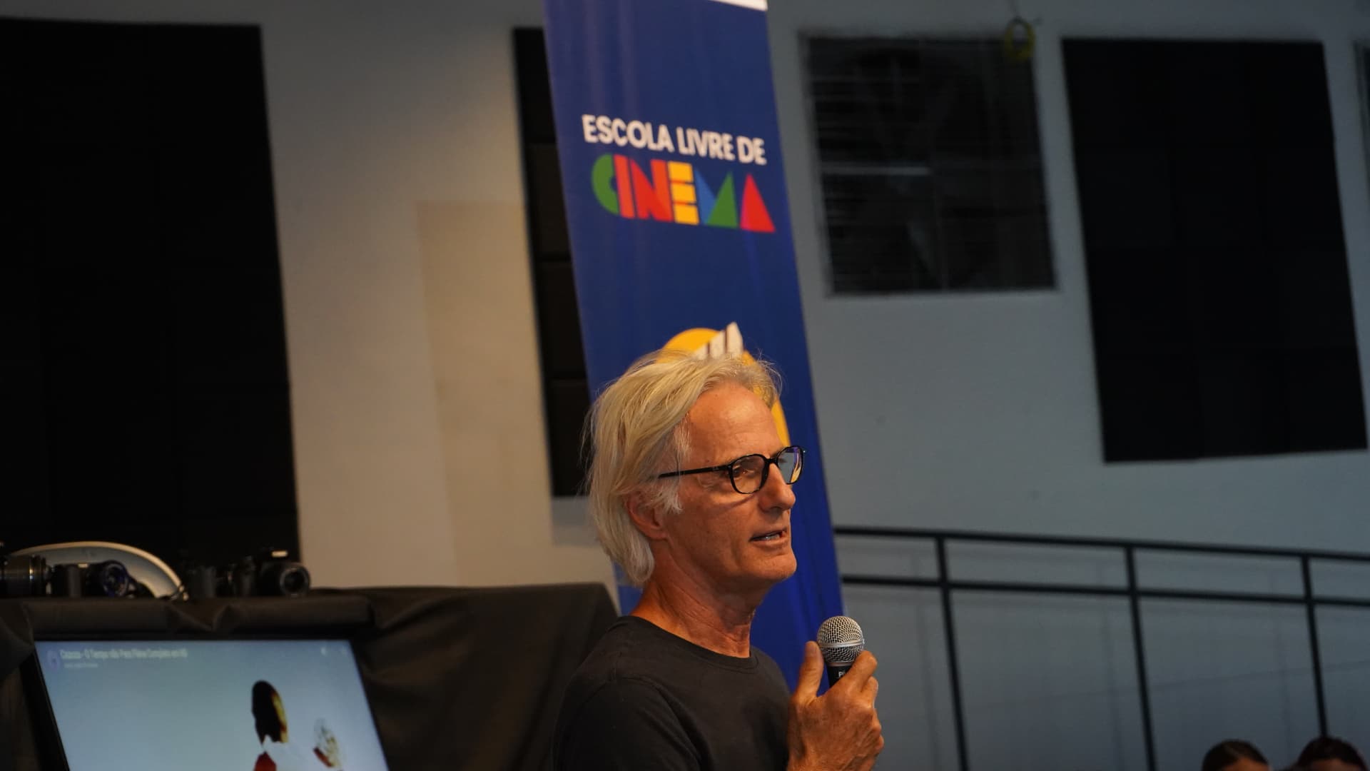 Pessoa com microfone diante do banner da Escola Livre de Cinema, em Extrema (MG), com câmeras e lentes sobre uma mesa e a projeção de um filme em um monitor